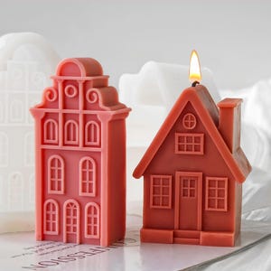 May include: Two house-shaped candles in shades of terracotta and coral. The taller candle features a stepped gable facade with multiple arched windows. The shorter candle is shaped like a cottage with a peaked roof, a chimney, and a lit wick.