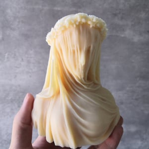May include: A cream-colored bust sculpture with a draped veil. The sculpture features a detailed, textured veil that obscures the face, and a ruffled crown. The bust is held in a hand against a gray background.