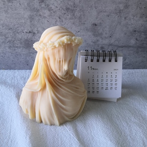 May include: A cream-colored bust sculpture of a veiled woman, adorned with a floral crown. The sculpture is next to a small calendar showing the month of November 2021. The sculpture's draped veil obscures the face, creating a mysterious aesthetic.