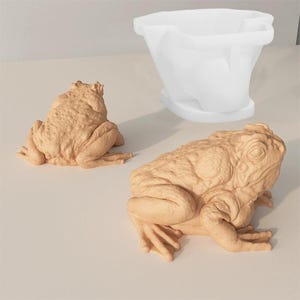 May include: Two tan-colored frog sculptures with detailed skin textures are displayed. One frog is in a sitting position, while the other is posed as if it is about to hop. A white, bowl-shaped object is in the background.