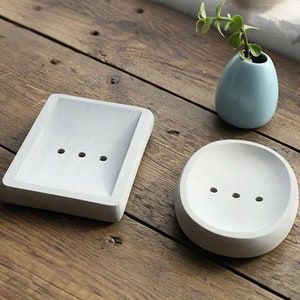 May include: Two white ceramic soap dishes, one square and one round, with drainage holes. The dishes are on a wooden surface.
