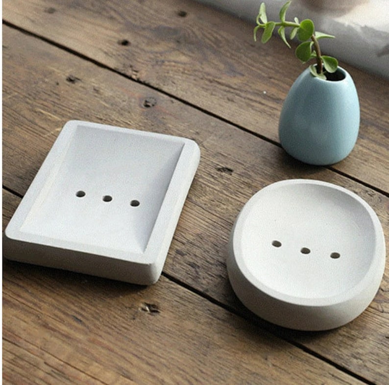 May include: Two white ceramic soap dishes, one square and one round, with drainage holes. The dishes are on a wooden surface.