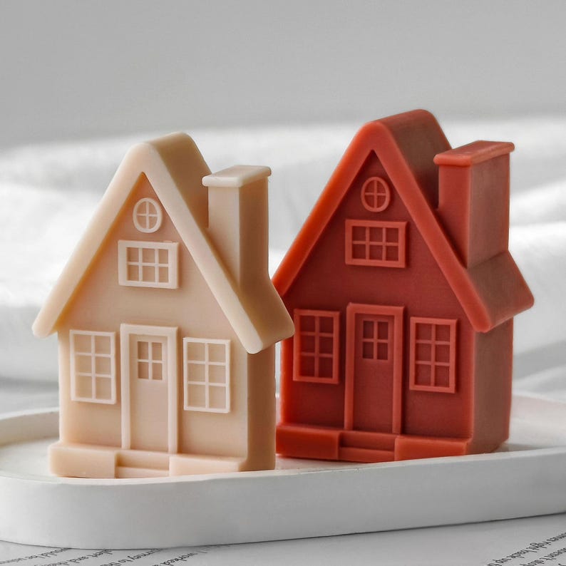 May include: Two house-shaped candles, one in a light beige and one in a deep terracotta red. Each candle features a molded design with a central door, multiple grid-patterned windows, a peaked roof, and a chimney on the side, all set on a tray.