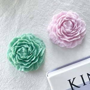 May include: Two floral-shaped soaps. One is a soft green, the other a pale pink. Both soaps have detailed petal designs. The soaps are on a white textured surface. A book with the word "KIN" is partially visible.