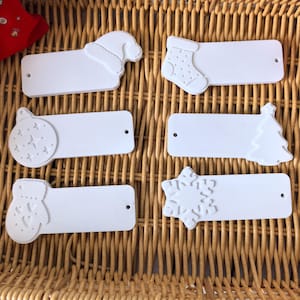 May include: Six white Christmas-themed gift tags with embossed designs. The tags feature a Santa hat, stocking, ornament, Christmas tree, mitten, and snowflake. Each tag has a small hole for stringing. The tags are arranged on a woven wicker surface.
