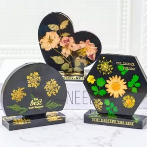 May include: Three black resin paperweights with different shapes and dried flower designs. The heart-shaped paperweight has pink and white flowers. The round paperweight has yellow flowers. The hexagonal paperweight has yellow and green flowers. The paperweights have the text "You deserve the best" on the base.