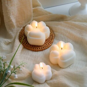 May include: Three white paw-shaped candles with a flame burning in each. The candles are on a woven wicker coaster and a white fabric background.
