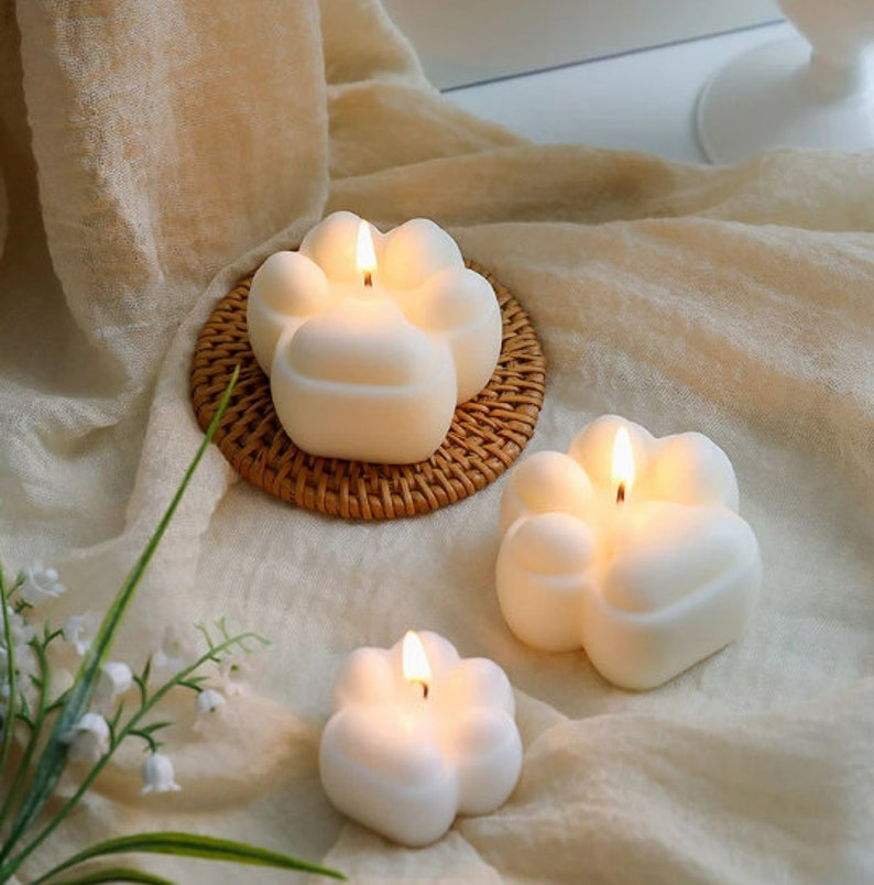 May include: Three white paw-shaped candles with a flame burning in each. The candles are on a woven wicker coaster and a white fabric background.
