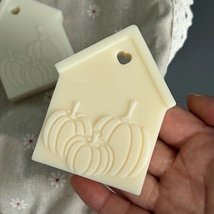 May include: An off-white wax melt shaped like a house with a small heart-shaped cutout near the roof peak. The front surface features a raised, embossed design of three pumpkins. A second identical wax melt is visible in the blurred background.