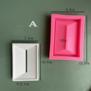 May include: A white rectangular soap dish with a raised center and a narrow slot. The dish measures 7.4 inches wide, 10.6 inches long, and 2.1 inches high. A pink silicone mold of the same shape is shown next to it.