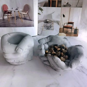 Op de afbeelding: Twee grijze betonnen handvormige potten. De ene pot is gevormd als een gesloten vuist en de andere is gevormd als een open hand die kleine stenen vasthoudt.