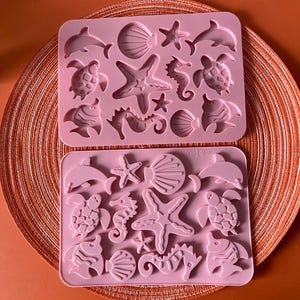 Könnte beinhalten: Zwei rosa Silikonformen mit verschiedenen Meeresbewohnern. Die Formen umfassen Delfine, Seesterne, Muscheln, Seepferdchen, Schildkröten und Fische. Die Formen sind auf einem orangefarbenen, gewebten Platzset angeordnet.