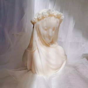 May include: A cream-colored candle shaped like a veiled bust. The candle features a floral crown and draped fabric, creating a serene and elegant aesthetic. The bust is a soft, off-white color.