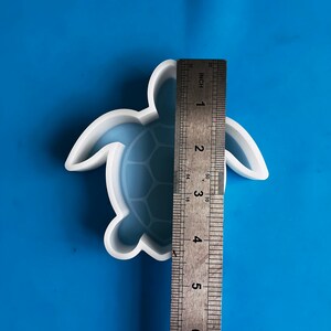 Puede incluir: Un molde blanco y azul con forma de tortuga, de aproximadamente 13 cm de largo, se muestra sobre un fondo azul. El molde tiene un dise&ntilde;o detallado de caparaz&oacute;n de tortuga. Se coloca una regla junto al molde para comparar el tama&ntilde;o.
