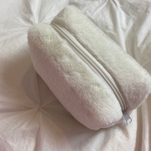 Peut inclure: Une trousse blanche et duveteuse avec une fermeture éclair argentée. L'étui rectangulaire est fait de fausse fourrure douce. La fermeture éclair longe le dessus pour l'ouverture et la fermeture.