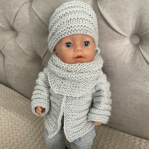 Peut inclure: Une tenue de poupée en tricot gris avec un chapeau, une écharpe et un cardigan. La poupée a les yeux bleus et porte un pantalon gris.