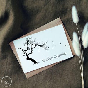 Puede incluir: Una tarjeta de felicitación rectangular blanca con una ilustración de silueta negra de un árbol desnudo con hojas que vuelan con el viento. El texto In stillem Gedenken está impreso en negro en la parte inferior derecha de la tarjeta, que descansa sobre un sobre marrón.