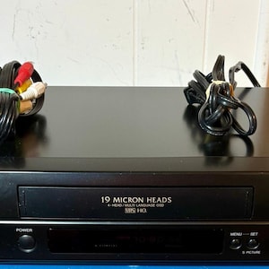 May include: A black Sharp VC-A552 VHS player featuring a front-loading cassette slot labeled 19 Micron Heads and VHS HQ. The device includes a circular control dial for play, pause, still, and eject functions, along with power, menu, and set buttons.