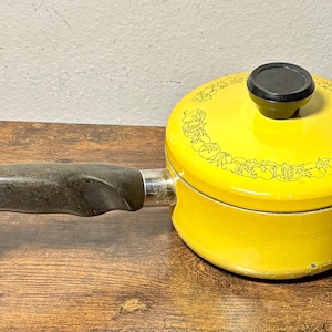 May include: A yellow saucepan with a black handle and lid. The lid has a black knob and a decorative border. The saucepan is sitting on a wooden surface.