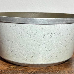 May include: A speckled, light gray cooking pot with a silver rim and handles. The pot has a wide opening and a rounded base. The interior is a darker gray. The pot is sitting on a wooden surface.