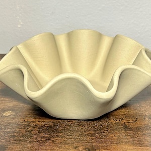 May include: A beige ceramic bowl with a ruffled edge. The bowl features a wavy, scalloped rim and a shallow, open design. The neutral colour and unique shape make it a decorative piece for the home.