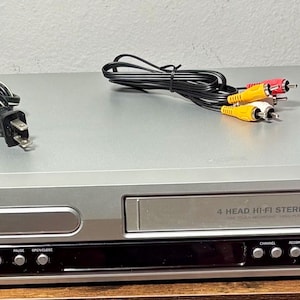 May include: Silver Magnavox DVD/VCR combo player with a black control panel and a VCR slot. The player has a power cord and a set of red, yellow, and white RCA cables. The unit is on a wooden surface.