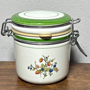 Pode incluir: Um frasco de cerâmica branco com tampa verde e fecho de metal. O frasco apresenta um desenho floral com morangos e mirtilos. A tampa tem uma borda verde. O frasco foi concebido para armazenamento.