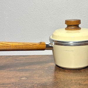 May include: A cream-colored saucepan with a lid and a wooden handle. The handle is attached to the pan with a metal bracket. The lid has a wooden knob. The pan is sitting on a wooden surface.