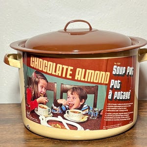 May include: Vintage soup pot with a brown lid and handles. The pot features a label with the words "Chocolate Almond" and an illustration of two children eating soup. The pot is beige with brown accents.