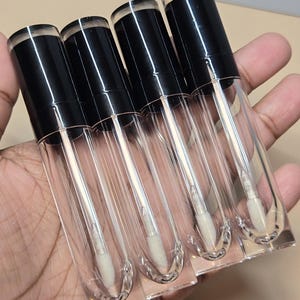 May include: A collection of five clear lip gloss tubes with black caps. Each tube has a clear applicator wand with a white, fluffy tip. The tubes are held in a hand, showcasing their transparent design.