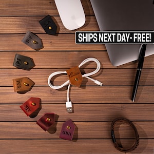 May include: A set of leather cord organizers with various initials and a Starburst logo. The organizers are brown, black, red, and purple. The organizers are on a wooden surface with a white cord and a laptop.
