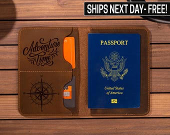 May include: Brown leather passport holder with a compass and the words "Adventure Time" engraved on the front. The holder has slots for cards and a pocket for a passport. The passport holder is open and a US passport is visible inside.
