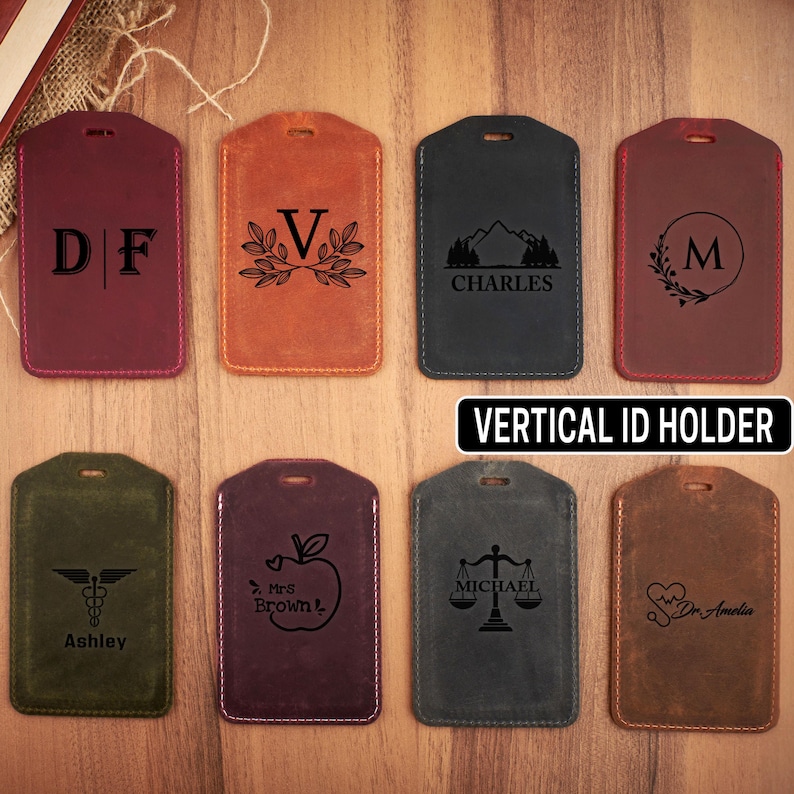 May include: Eight different leather vertical ID holders with various designs and text. The designs include a mountain range, a heart with a stethoscope, a scale of justice, an apple with a teacher's name, a caduceus, a wreath with a letter V, a wreath with a letter M, and initials D and F.