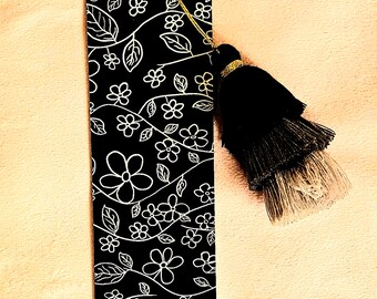 May include: Black bookmark with a white floral pattern and a black tassel. The bookmark features a repeating design of flowers and leaves. A gold string and bead attach the tassel to the bookmark.