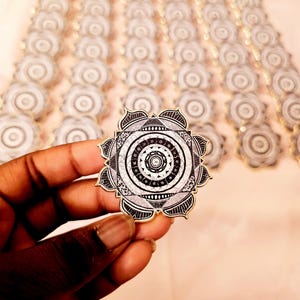 Peut inclure: Épinglette mandala noire et blanche avec des motifs géométriques complexes, tenue dans une main. L'épinglette a une bordure dorée et un design détaillé. De nombreuses épinglettes similaires sont disposées en rangées en arrière-plan.