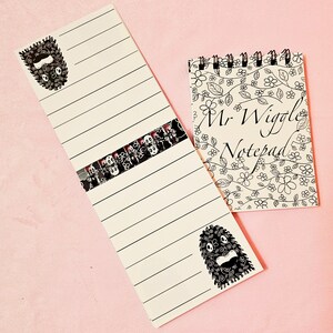 Peut inclure: Un bloc-notes et un long bloc-notes ligné avec des illustrations en noir et blanc sur fond rose. Le bloc-notes indique "Mr. Wiggle Notepad" et possède une bordure florale. Le long bloc-notes a une bande décorative de têtes de mort.