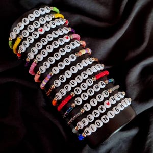 May include: A stack of colorful beaded bracelets with white letter beads spelling out the names of Taylor Swift albums, including "Folklore", "Debut", "Fearless", "Lover", "1989", "Reputation", "Red", "Speak Now", and "Midnights".
