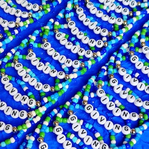 May include: A close-up of several colorful beaded bracelets with the word "GIVING" spelled out on each one. The bracelets are made with blue, green, and white beads and silver spacers.
