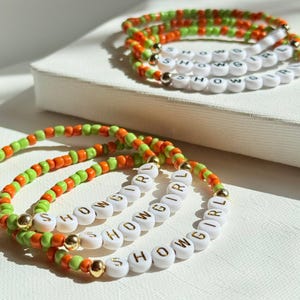 May include: Several beaded bracelets with the word "SHOWGIRL" spelled out in white letter beads. The bracelets feature orange and green beads, with gold-colored accents. The bracelets are displayed on a white surface.