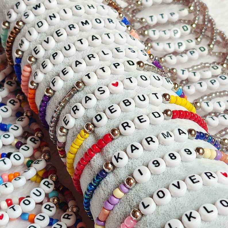 May include: A close-up of a stack of colorful beaded bracelets with white letter beads spelling out words like "Eras", "Debut", "Karma", "Lover", and "Tour".