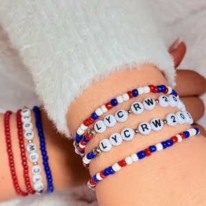 May include: A set of beaded bracelets featuring red, white, and blue seed beads. Two bracelets include white round letter beads spelling LYC RW 2021, interspersed with small silver-toned metallic spacer beads. The design uses a repeating color pattern.