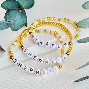 May include: Three beaded bracelets featuring alternating gold and white seed beads. Each bracelet includes white spherical beads with gold lettering spelling out the names MIKAYLA, SHERREYL, and SHERREYL, accented with small gold metallic spacer beads.