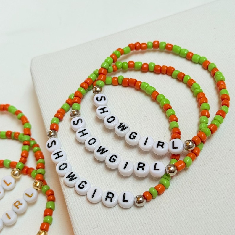 May include: Three beaded bracelets with the word "SHOWGIRL" spelled out in white letter beads. The bracelets feature orange and green beads, with gold-colored accents. The bracelets are on a white surface.