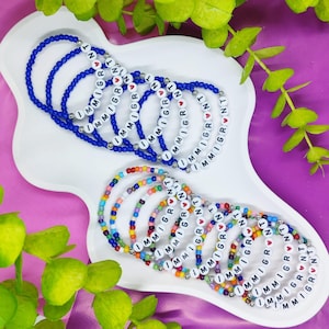 May include: A collection of elastic bracelets featuring white letter beads spelling the word IMMIGRANT, interspersed with small silver-toned spacer beads. One set consists of blue beads, while the other set features a multicolored bead pattern.