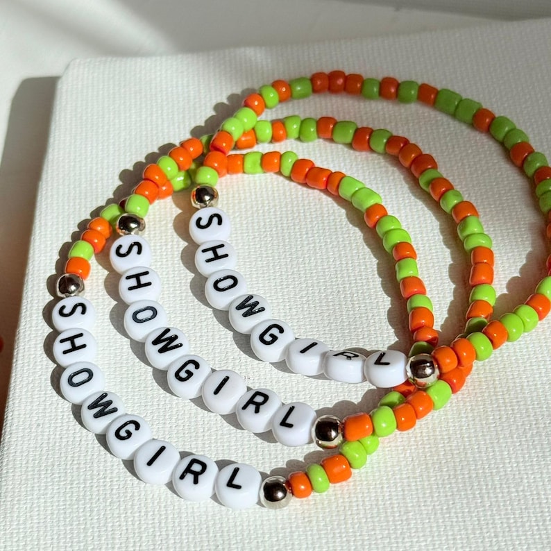 May include: Three beaded bracelets with the word "SHOWGIRL" spelled out in white letter beads. The bracelets feature orange, green, and gold beads. The bracelets are on a white surface.