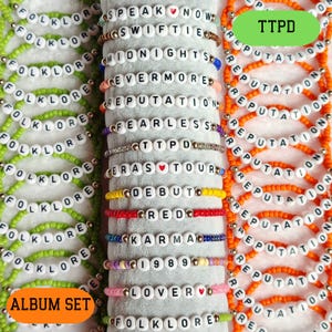 May include: A set of 18 beaded bracelets with the names of Taylor Swift's albums. The bracelets are in green, orange, red, yellow, blue, purple, and pink. The bracelets are arranged in two rows, with the album names "Folklore" and "Reputation" at the top of each row. The text "TTPD" is visible in the top right corner of the image.