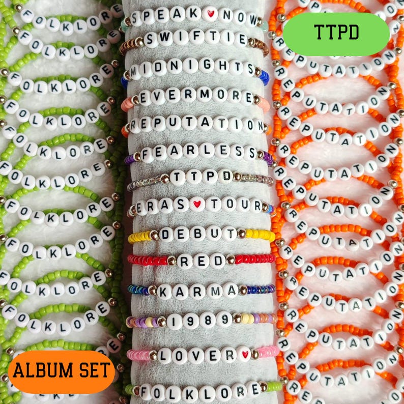 May include: A set of 18 beaded bracelets with the names of Taylor Swift's albums. The bracelets are in green, orange, red, yellow, blue, purple, and pink. The bracelets are arranged in two rows, with the album names "Folklore" and "Reputation" at the top of each row. The text "TTPD" is visible in the top right corner of the image.