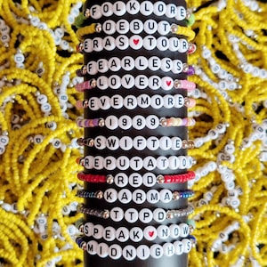 May include: A stack of colorful beaded bracelets with white letter beads spelling out the names of Taylor Swift's albums, including "Folklore", "Debut", "Eras Tour", "Fearless", "Lover", "Evermore", "1989", "Speak Now", and "Midnights".