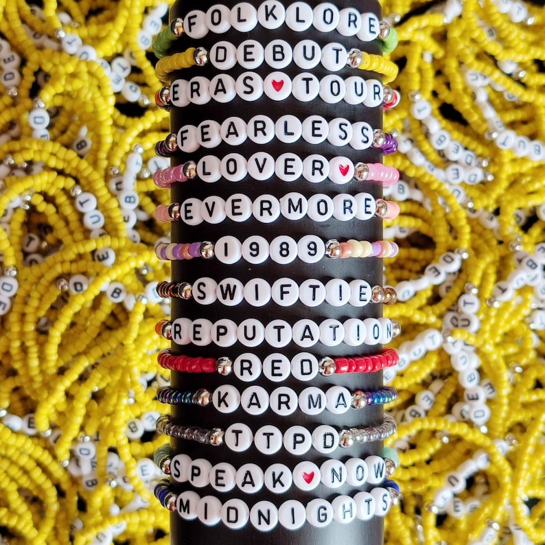 May include: A stack of colorful beaded bracelets with white letter beads spelling out the names of Taylor Swift's albums, including "Folklore", "Debut", "Eras Tour", "Fearless", "Lover", "Evermore", "1989", "Speak Now", and "Midnights".