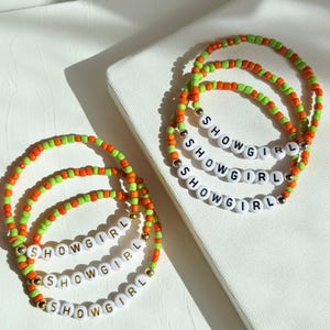 May include: Three beaded bracelets with the word "SHOWGIRL" spelled out in white letter beads. The bracelets feature a combination of orange, green, and gold beads. The bracelets are displayed on a white surface.